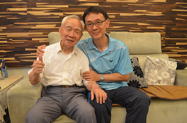 老爺爺和兒子一起坐在綠色沙發上，開心的合照，爺爺手比讚