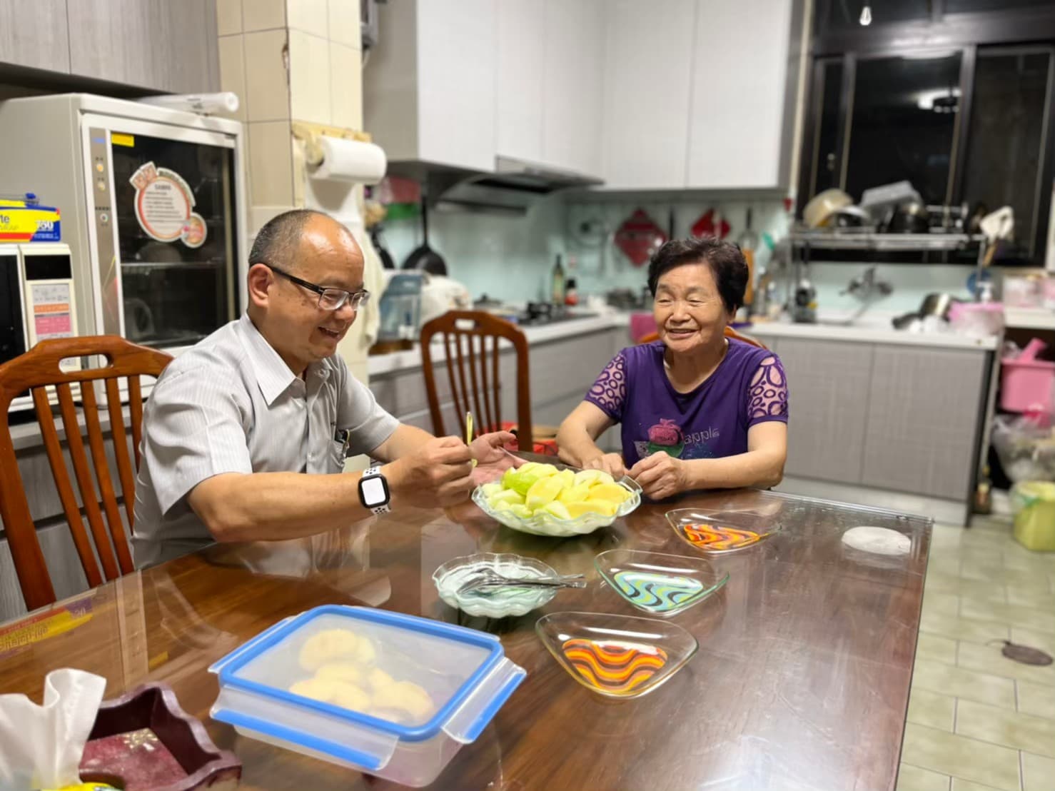 男子與老母親坐在廚房裡邊吃水果邊聊天