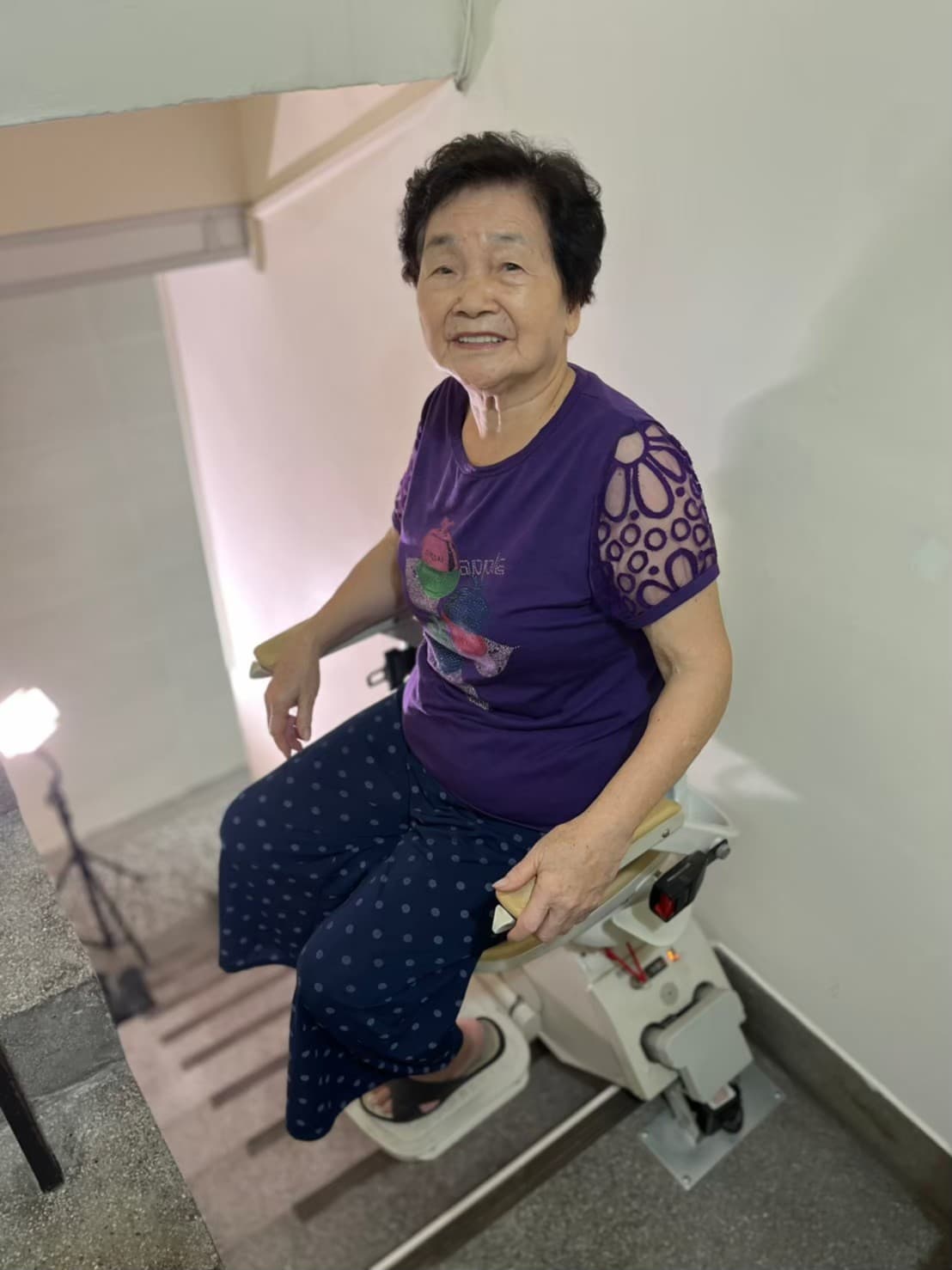 身穿紫色衣服的黑髮奶奶坐在泓電樓梯升降椅上微笑看著鏡頭